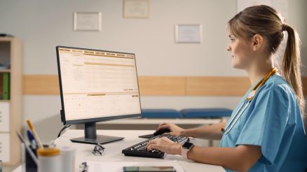 Junge Ärztin am Schreibtisch mit Stethoskop um den Hals arbeitet am Computer