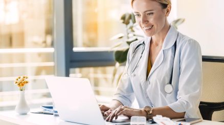 Eine Ärztin mit Stethoskop um den Hals sitzt am Schreibtisch und arbeitet am Laptop