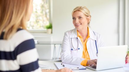 Ärztin sitzt am Schreibtisch vor einem Laptop und ist im Gespräch mit einer Patienten