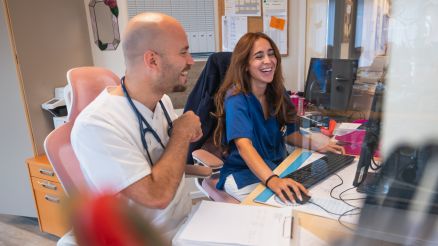 Zwei Krankenpfleger*innen sitzen vor einem PC und lachen